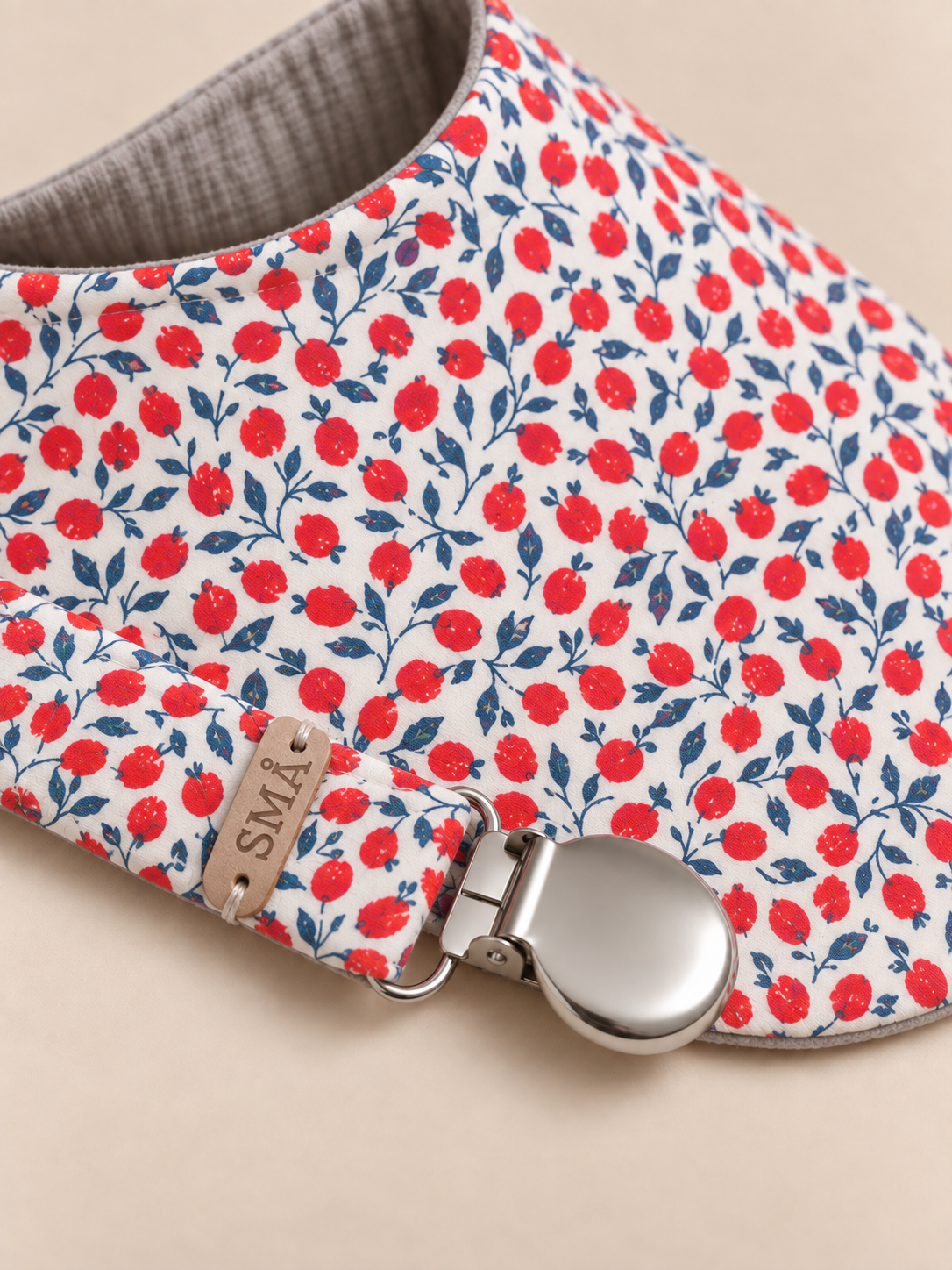 Close-up detail of SMÅ baby bib and pacifier clip featuring red berry and navy botanical print, grey textured lining and silver clip hardware