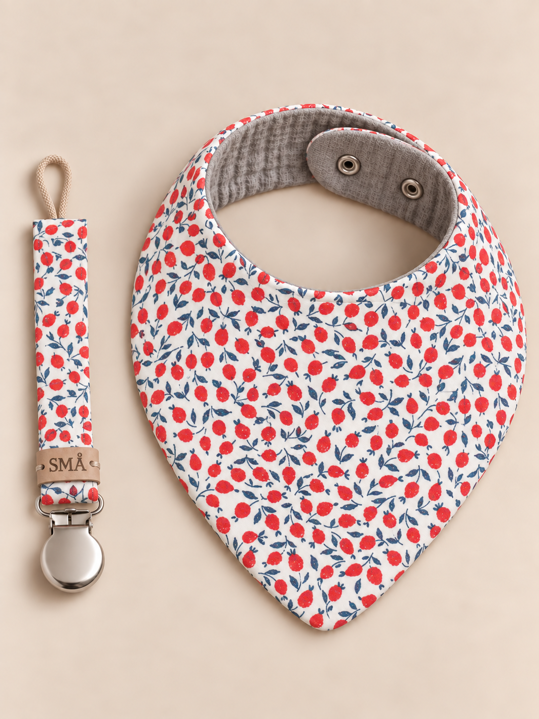 SMÅ baby bib and matching pacifier clip in cream fabric with red berry and navy leaf print, photographed on a neutral beige background
