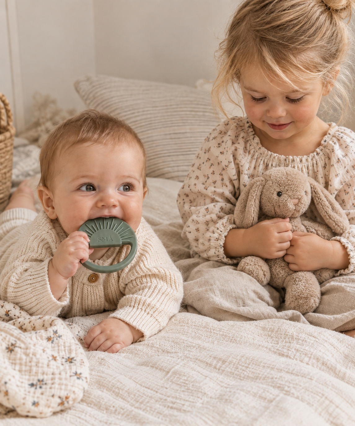 Baby holding and chewing a sage green SMÅ Sun Baby Teether while an older girl sits beside them with a soft crochet bunny on a cozy bed in neutral tones