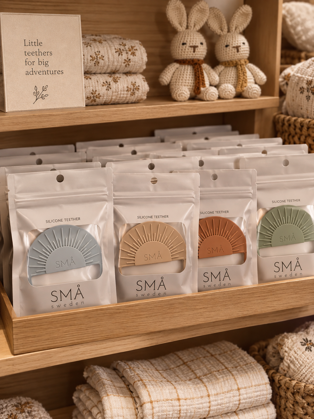 SMÅ Sun Baby Teethers in light gray, sand, caramel and eucalyptus displayed in branded packaging on a wooden boutique shelf with crocheted rabbits and folded muslin swaddles