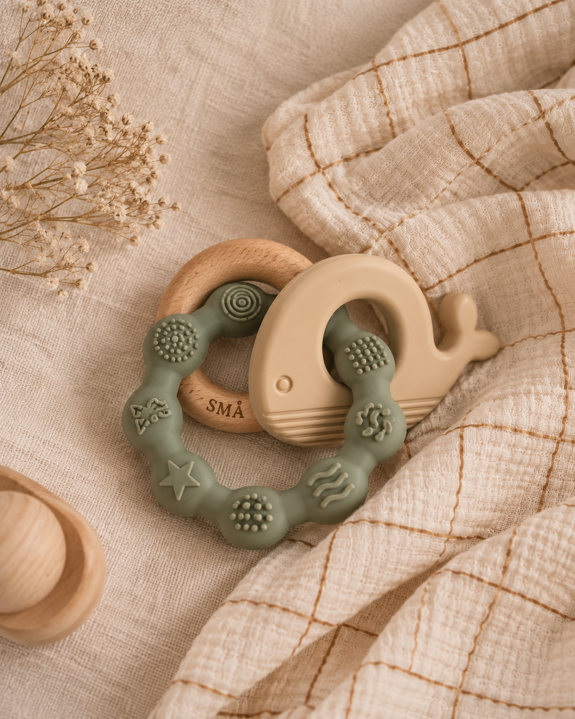 SMÅ whale baby teether in sage green and natural wood, styled on soft cream linen with a glimpse of the brand’s checked muslin swaddle.