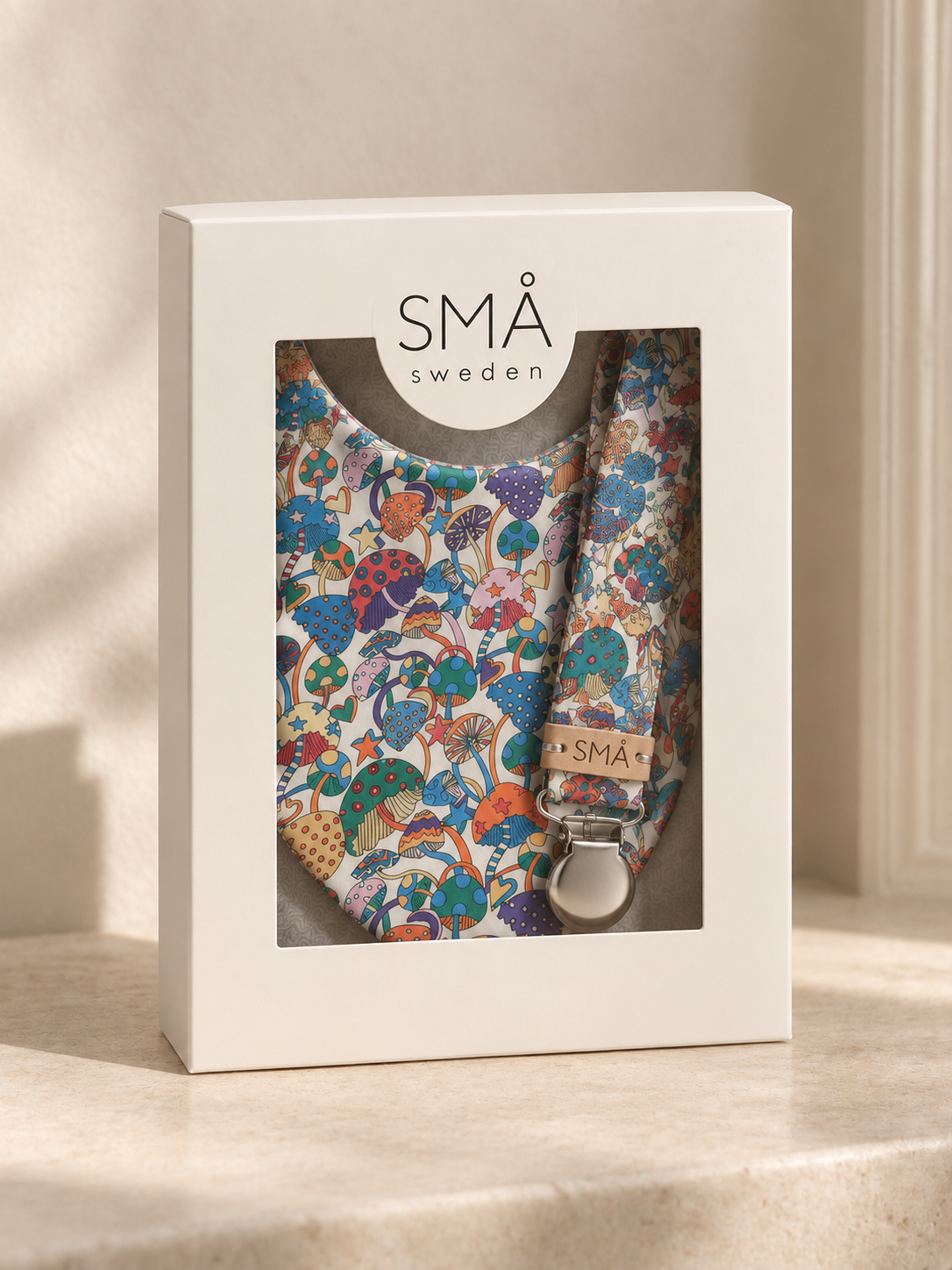 White SMÅ Sweden gift box with clear window containing Liberty Magic baby bib and matching pacifier clip