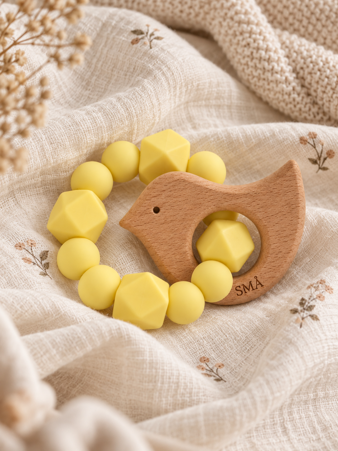 SMÅ Birdy Baby Teether in yellow silicone and natural beech wood resting on floral muslin fabric