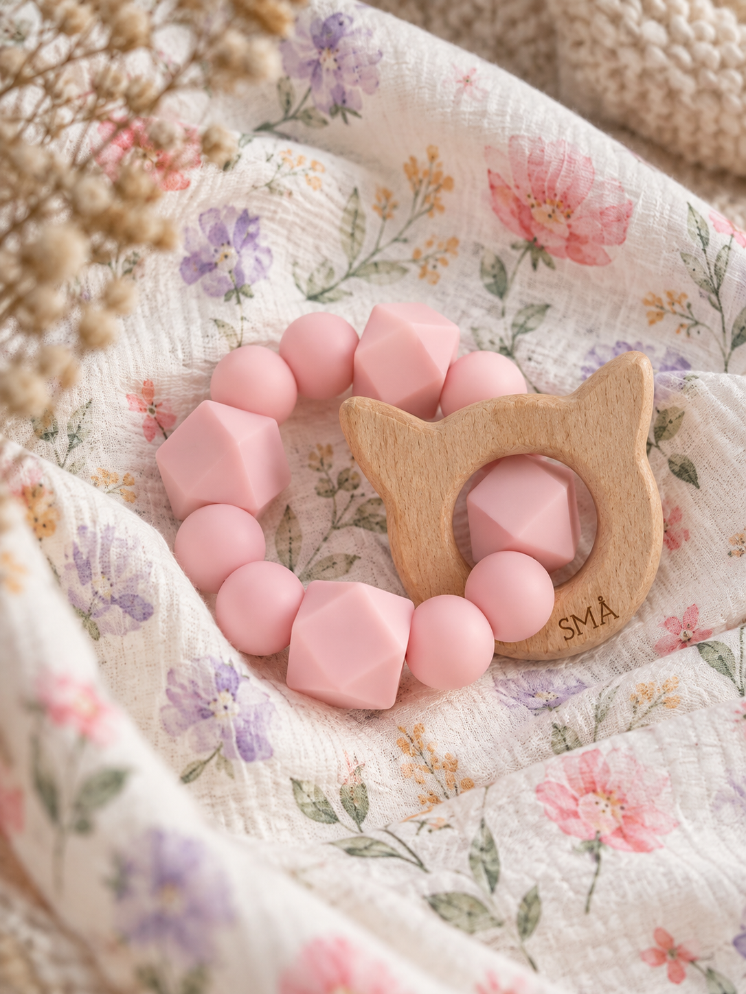SMÅ Kitty Baby Teether in pink silicone and beech wood resting on floral muslin fabric