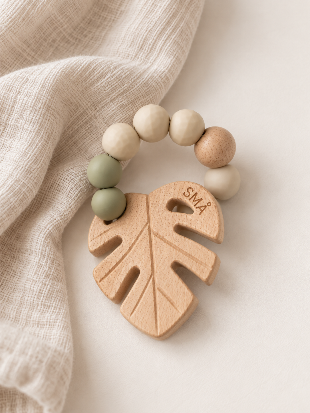 SMÅ Leaf Baby Teether in natural beech wood and sage green silicone arranged on a warm ivory background