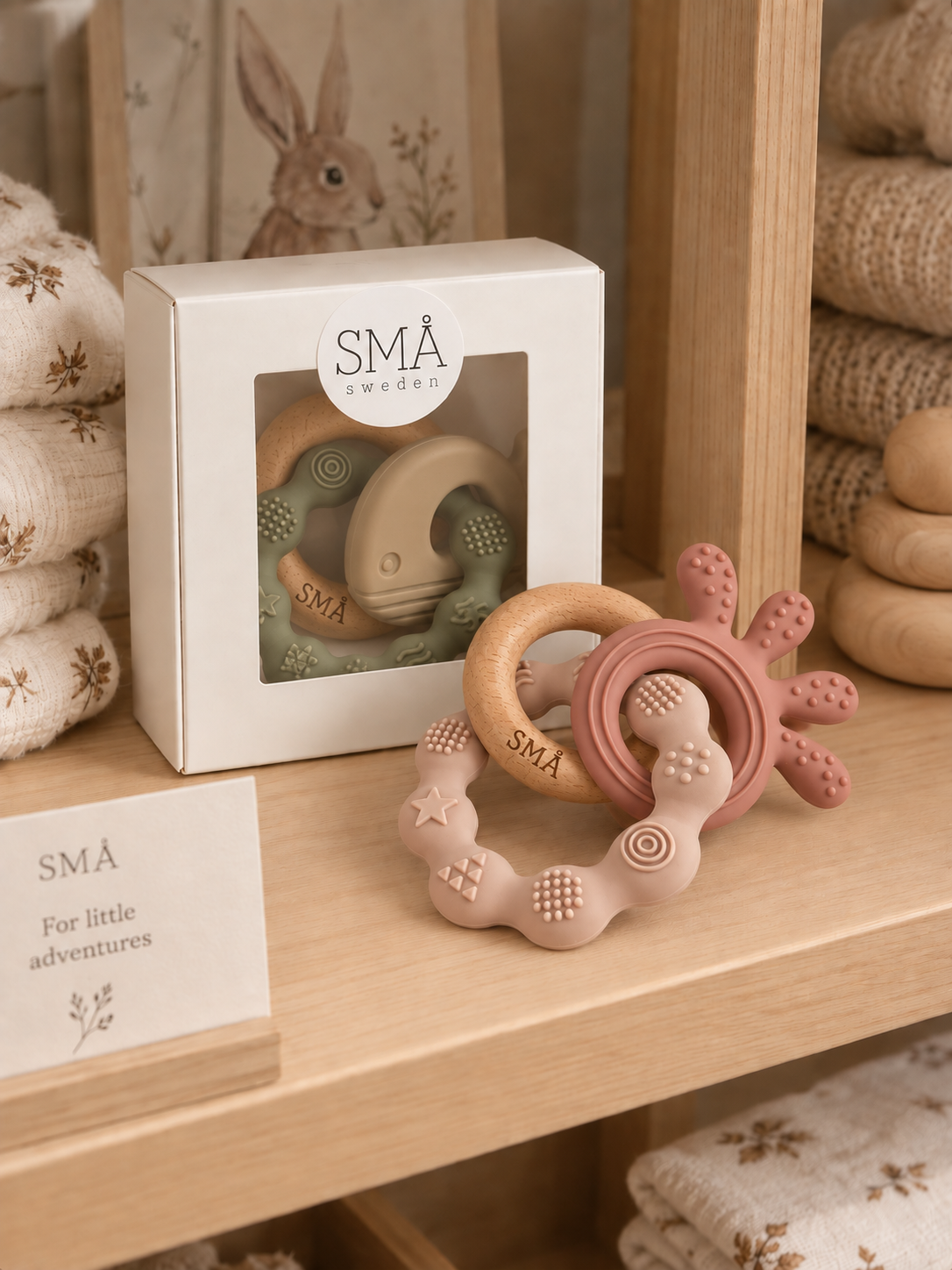 A set of SMA Sweet baby teething toys displayed next to their white packaging box on a wooden shelf, surrounded by styled baby accessories.